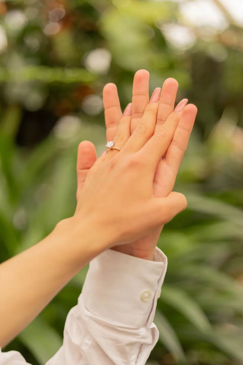 Hands showing an engagement ring against soft greenery.